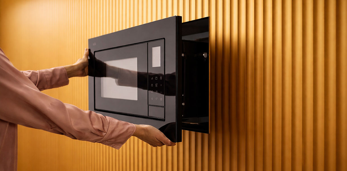 Handen die AEG 6000-serie inbouw magnetron in zwart openen tegen gouden geribbelde keukenachtergrond