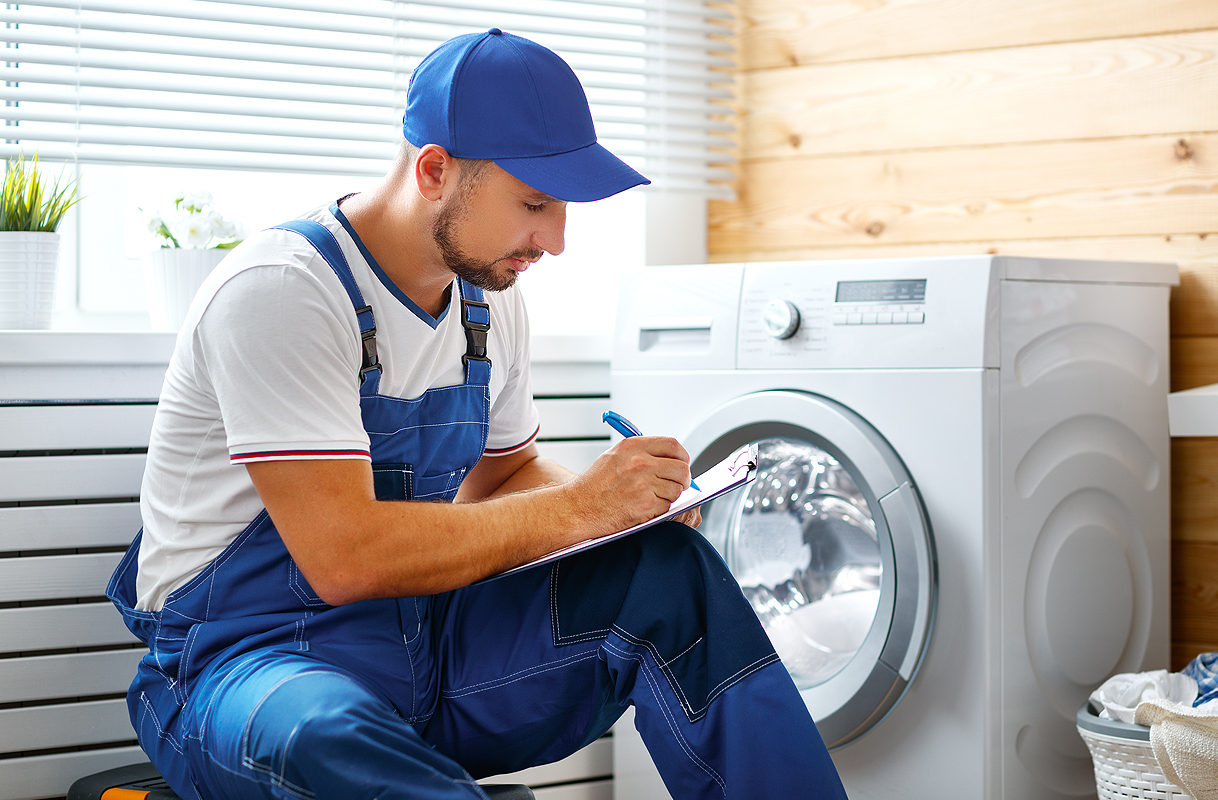 Monteur in blauwe overall hurkt naast een wasmachine en maakt aantekeningen op een klembord
