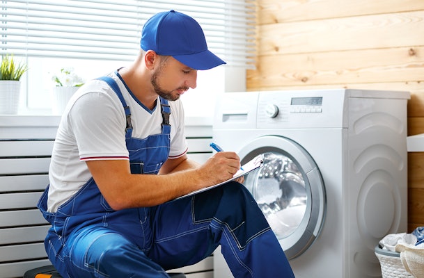 Monteur in blauwe overall hurkt naast een wasmachine en maakt aantekeningen op een klembord