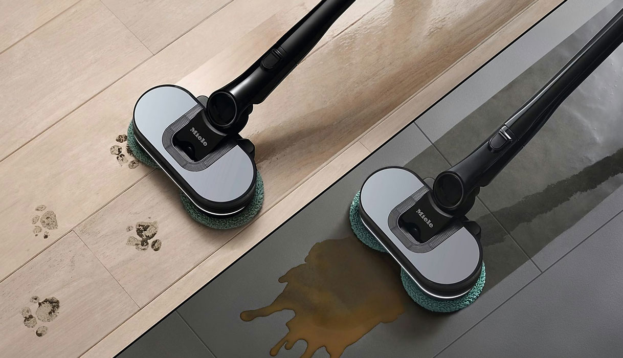 Twee Miele AquaTwister Pro dweilborstels reinigen modderige voetafdrukken op houten vloer en gemorste vloeistof op tegelvloer