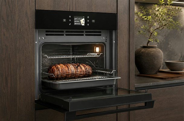 Bauknecht inbouw magnetron in een donker eiken keuken met open deur, rolade en kerntemperatuurmeter.