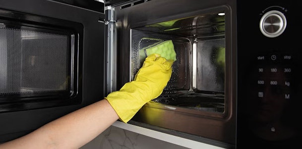 Handen met zwarte handschoenen die de binnenkant van een magnetron schoonmaken met een groene doek en sopje