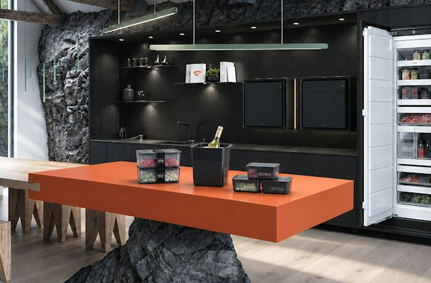 BORA Freeze inbouwvriezer met geopende deur en accessoires op een oranje kookeiland, waaronder voorraaddozen met diepvriesproducten, portievormen en een wijnkoeler.