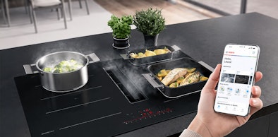 Smartphone met Home Connect app naast een Bosch kookplaat met afzuiging