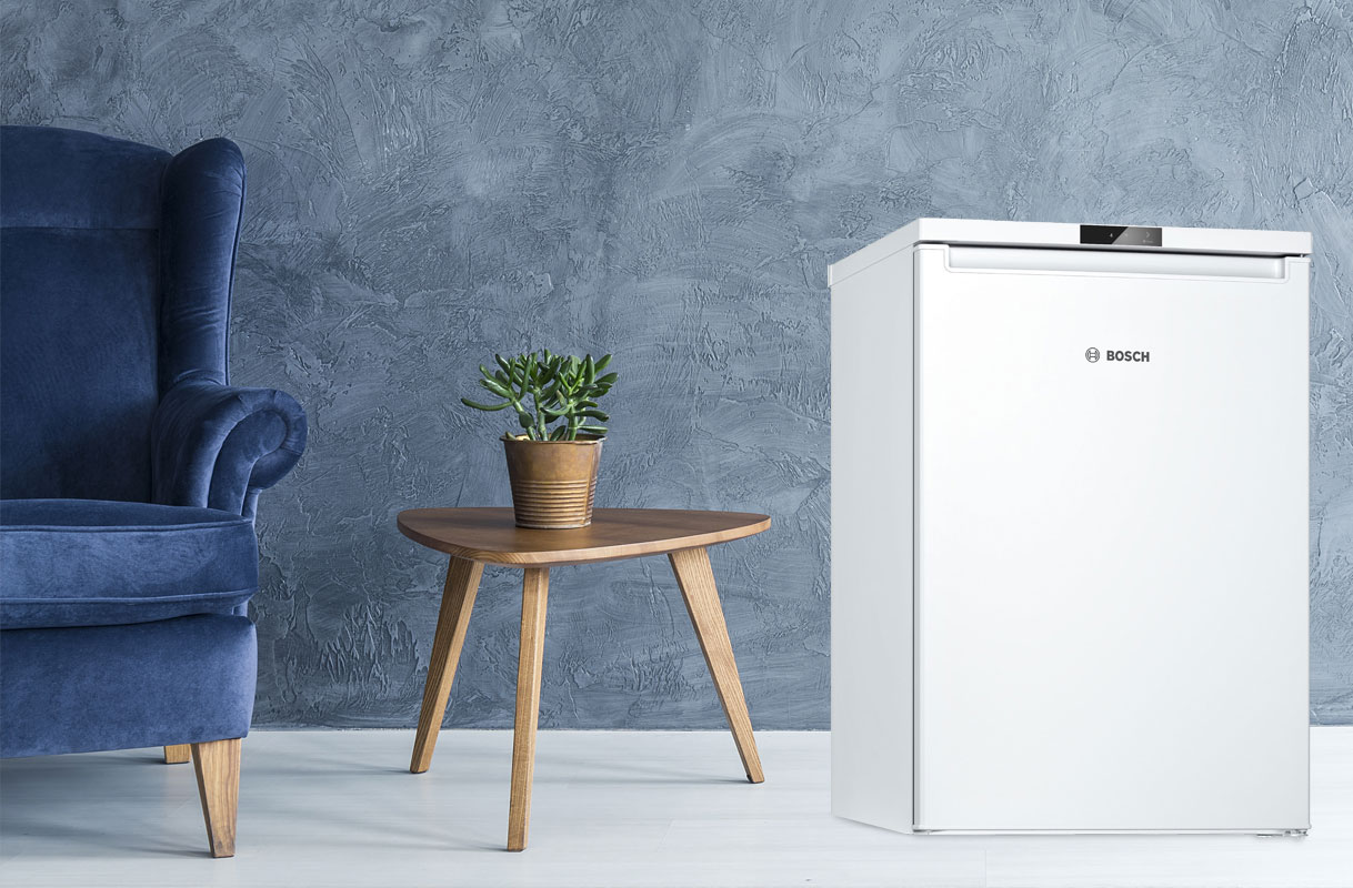 Bosch tafelmodel koelkast in witte uitvoering naast houten bijzettafel met plant en blauwe fauteuil in modern interieur.