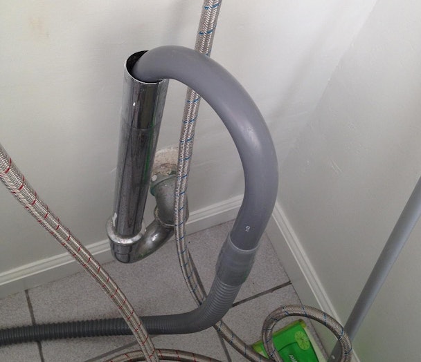 Grijze afvoerslang van wasmachine hangt in een chromen standpijp, met watertoevoerslangen en wandaansluiting zichtbaar