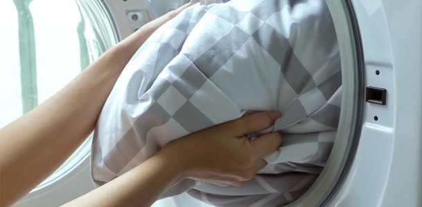 Handen duwen een wit-grijs geruit dekbed in de trommel van een wasmachine