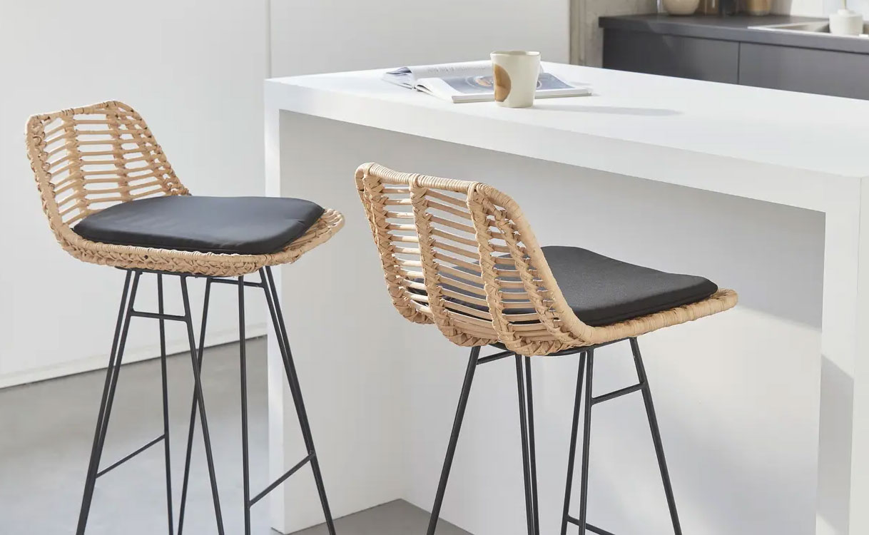 Twee design barkrukken met gevlochten rotan zitting en zwart kussen op slanke metalen poten naast een wit keukeneiland