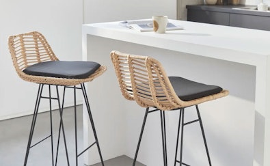Twee design barkrukken met gevlochten rotan zitting en zwart kussen op slanke metalen poten naast een wit keukeneiland