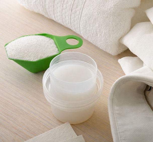 Waspoeder in groene maatschep en vloeibaar wasmiddel in maatbeker naast witte handdoeken