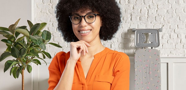 Vrouw met bril glimlacht in wasruimte met strijkplank op achtergrond