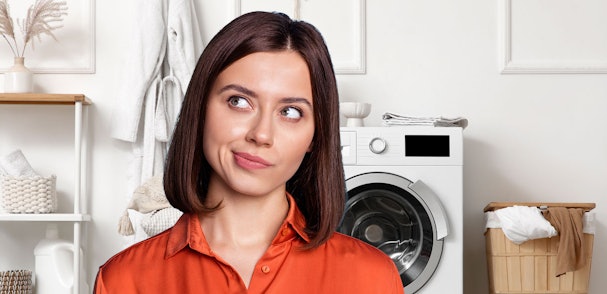 Vrouw met donker haar in oranje blouse kijkt nadenkend opzij, met een wasmachine en wasmand op de achtergrond