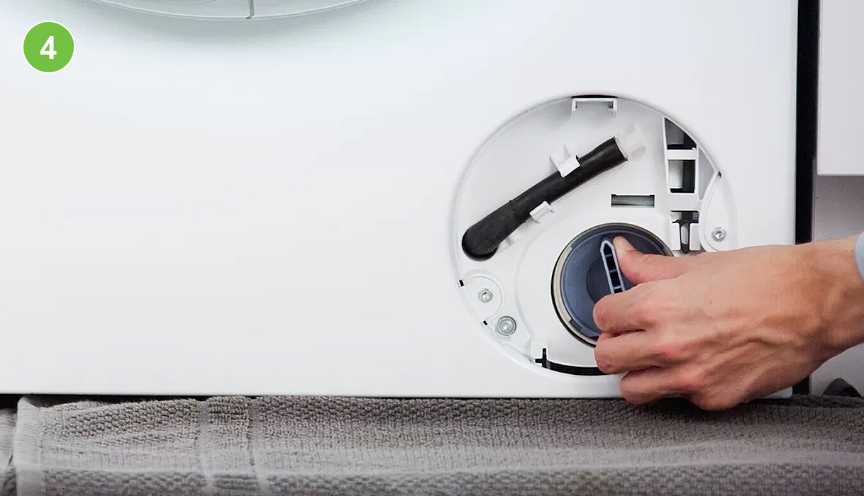 Hand draait het pompfilter van een wasmachine langzaam los, een handdoek ligt eronder om water op te vangen