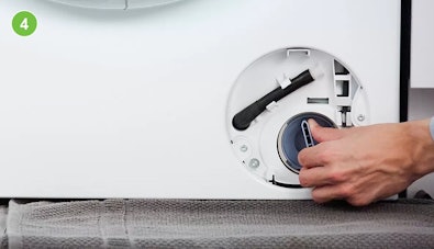 Hand draait het pompfilter van een wasmachine langzaam los, een handdoek ligt eronder om water op te vangen