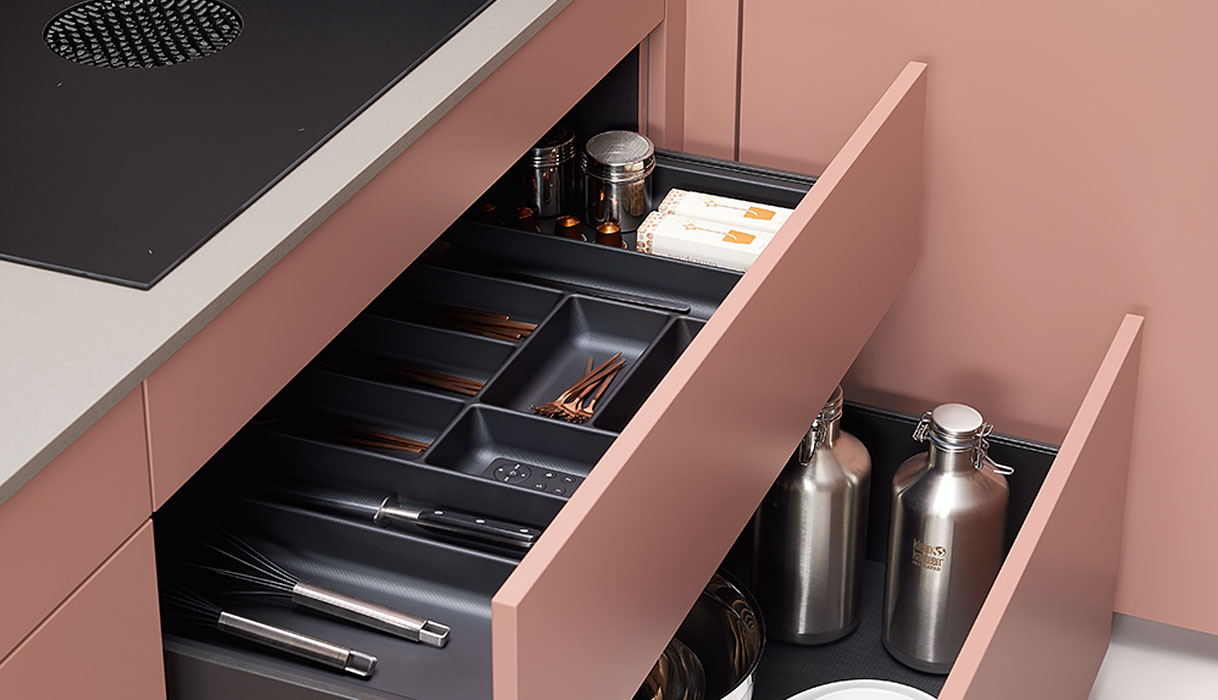 Zachtroze pastel keuken met geopende lade en bestekindeling onder zwarte inductiekookplaat.