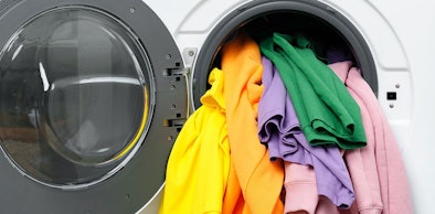 Alternatieve tekst (alt): Geopende wasmachine met kleurrijke kleding in geel, oranje, lila, groen en roze die uit de trommel hangt