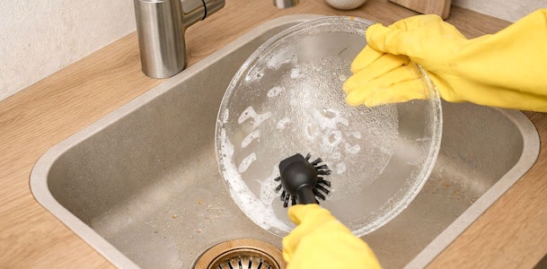 Handen met gele handschoenen die een glazen magnetronplaat schoonmaken met een borstel in de gootsteen