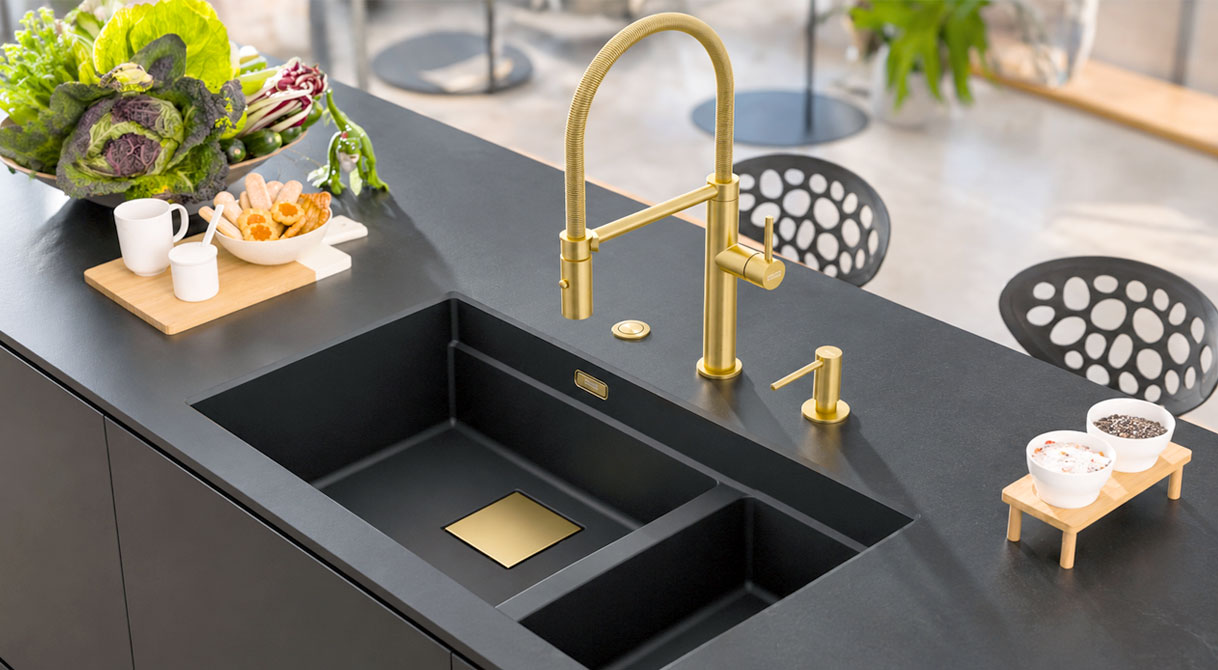 Gouden keukenkraan met flexibele uitloop en bijpassende zeepdispenser boven zwarte spoelbak in donker keukenblad