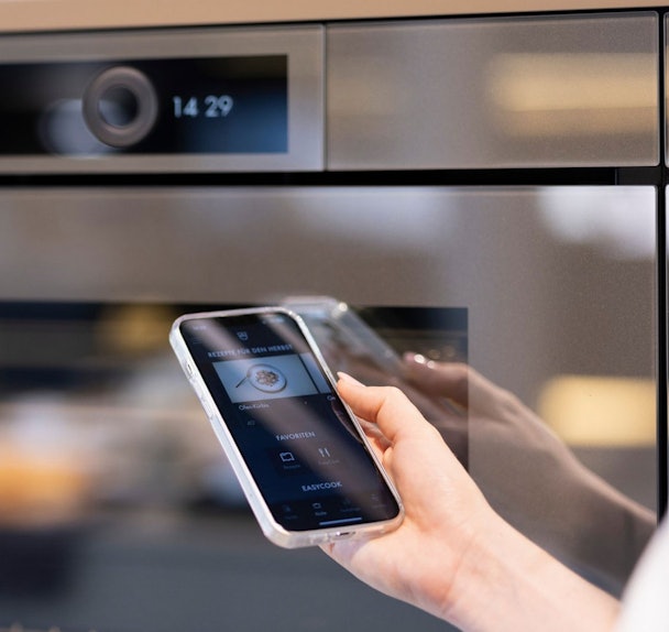 V-ZUG Home-app op smartphone gekoppeld aan keukenapparaat voor slimme bediening, recepten en persoonlijke instellingen.