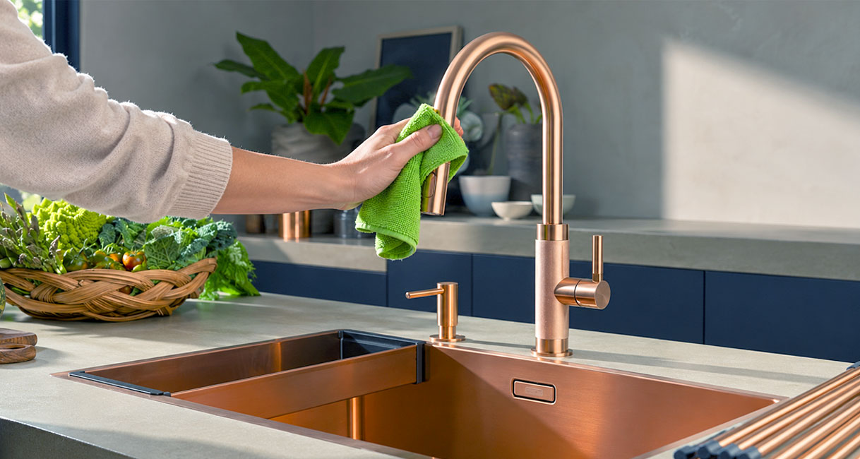 Hand maakt koperen keukenkraan schoon met groene doek boven koperen spoelbak