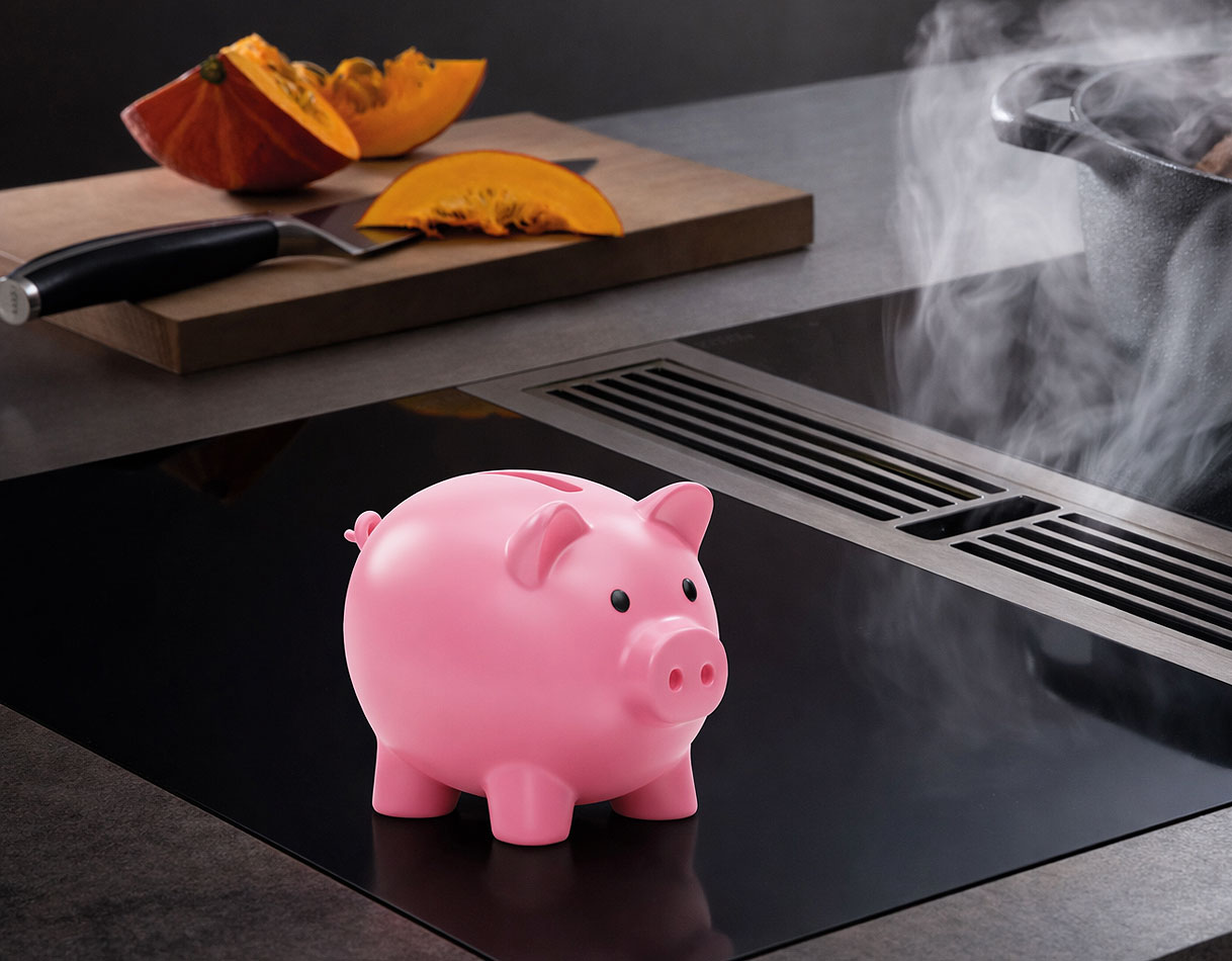 Inductiekookplaat met geïntegreerde afzuiging en een roze spaarvarken als symbool voor de kosten van overstappen van gas naar inductie.
