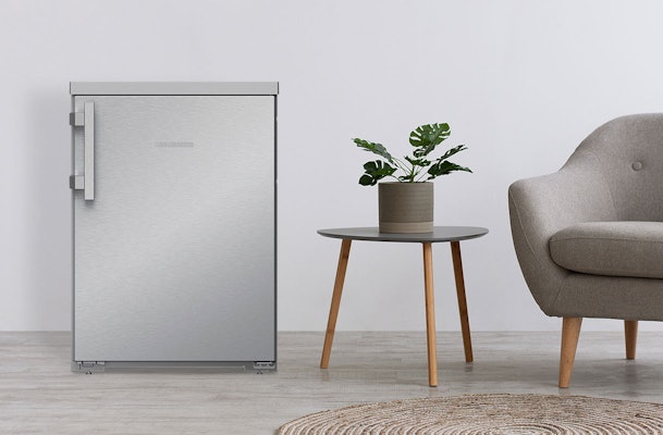 Liebherr tafelmodel koelkast in roestvrijstaal naast grijze stoel en bijzettafel met plant in modern interieur.
