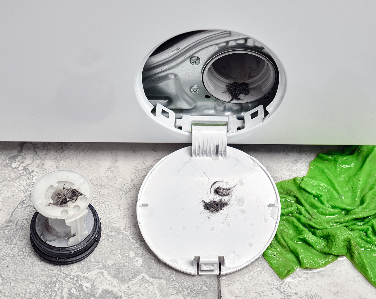 Geopend pompfilterluik van een wasmachine met verwijderd filter waarop haar en pluizen zichtbaar zijn, een groene handdoek ligt ernaast om water op te vangen
