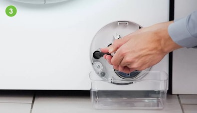 Alt-tekst (voor toegankelijkheid): Handen trekken de zwarte noodafvoerslang uit het filtercompartiment van een wasmachine, een doorzichtige bak staat eronder om het water op te vangen