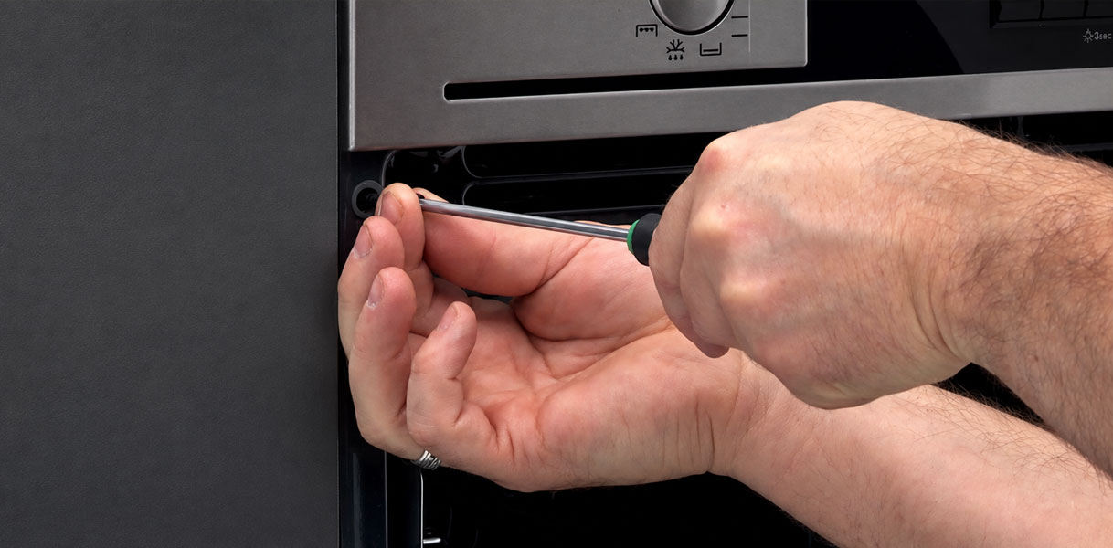 Hand draait met een schroevendraaier een bevestigingsschroef los in de deuropening van de oven