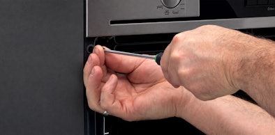 Hand draait met een schroevendraaier een bevestigingsschroef los in de deuropening van de oven