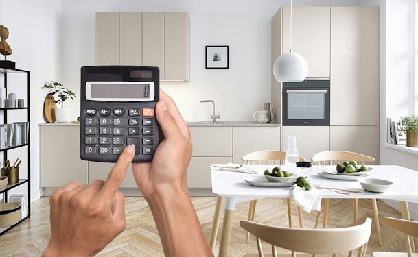 Persoon berekent de kosten van een moderne rechte keuken met calculator aan een lichte eetkeuken met beige fronten en compacte opstelling.