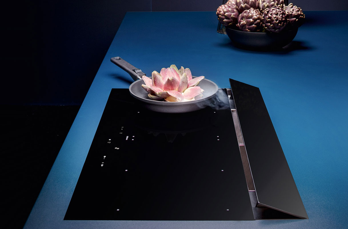 Falmec inductiekookplaat met geïntegreerde afzuiging en pan met stoom naast opklapbare afzuigklep.