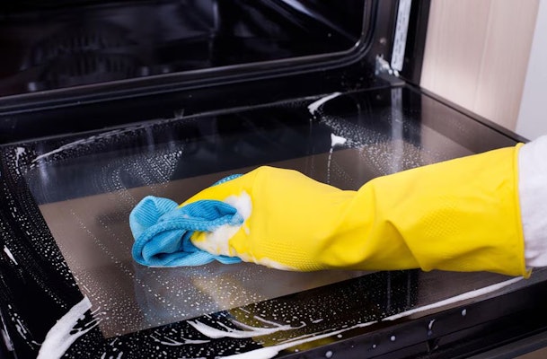 Hand met gele schoonmaakhandschoen veegt schuim weg van een ovenbodem met een zachte doek