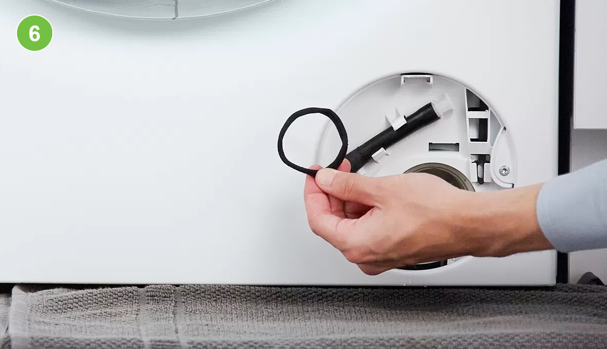 Hand houdt een haarbandje vast dat uit het pompfilter is gehaald, het filtercompartiment met pompschoep is zichtbaar op de achtergrond