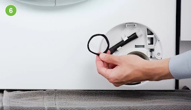 Hand houdt een haarbandje vast dat uit het pompfilter is gehaald, het filtercompartiment met pompschoep is zichtbaar op de achtergrond