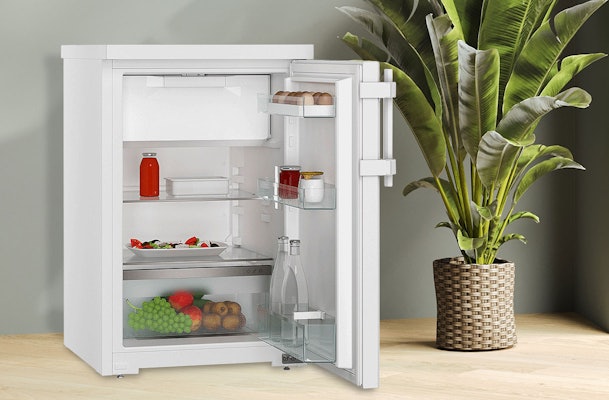 Vrijstaande tafelmodel koelkast met vriesvak, open deur met verse etenswaren en glazen plateaus, compact geplaatst.