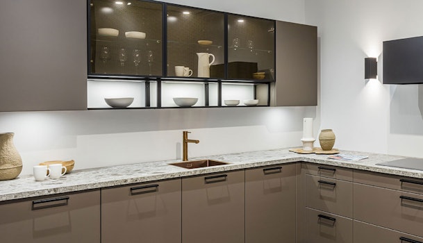 Taupe keuken met marmerlook werkblad, zwarte handgrepen en zwarte vitrinekasten met verlichting.