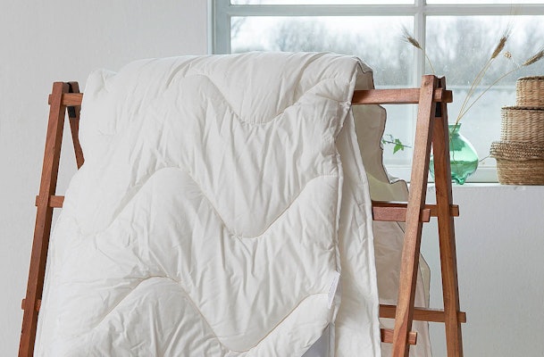 Wit dekbed hangt te luchten over houten kledingrek bij raam