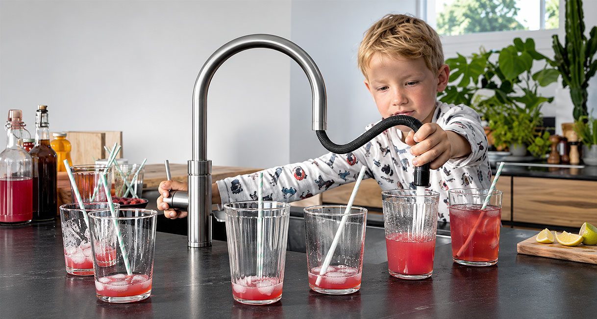 Kind trekt sproeikop met slang uit rvs keukenkraan om glazen limonade te vullen op het aanrecht