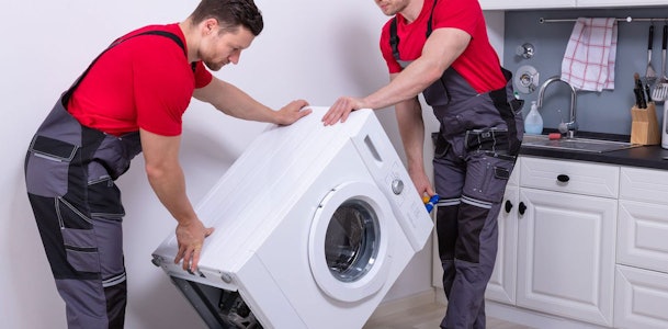 Twee monteurs tillen samen een wasmachine door deze schuin te kantelen