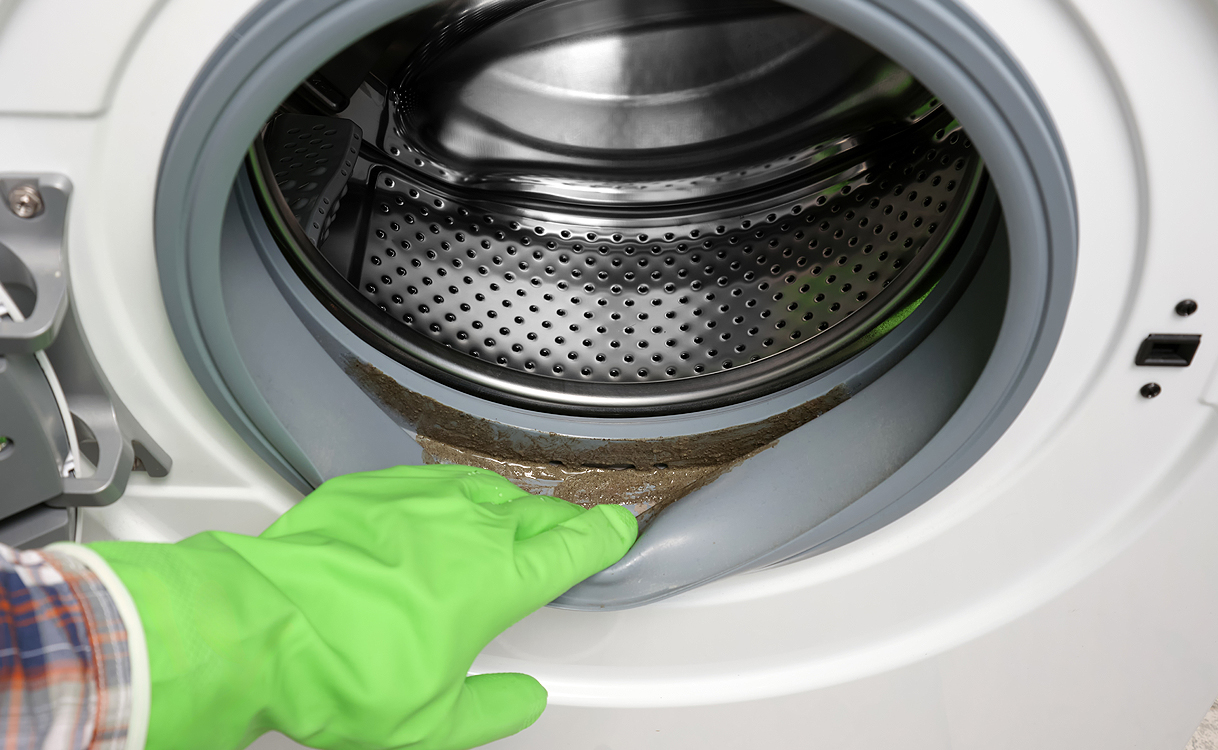 Hand met groene rubberen handschoen wijst naar grijsbruine vetluisaanslag op de manchet van een wasmachine
