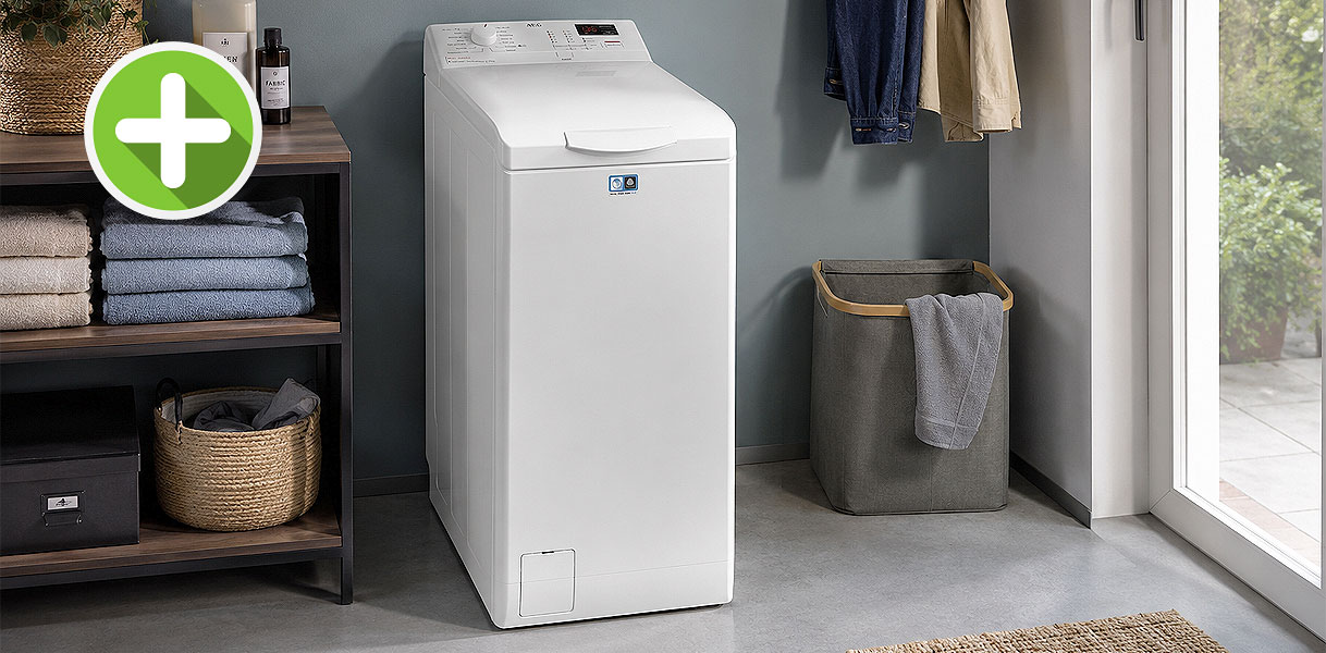 Smalle witte AEG bovenlader wasmachine in een bijkeuken naast een wasmand bij het raam