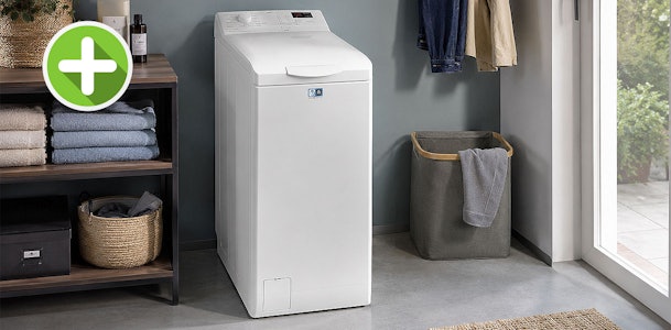 Smalle witte AEG bovenlader wasmachine in een bijkeuken naast een wasmand bij het raam