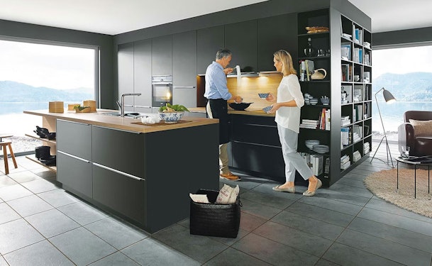 Een woonkeuken met een donker spoeleiland en een aangrenzende woonkamer met grote ramen