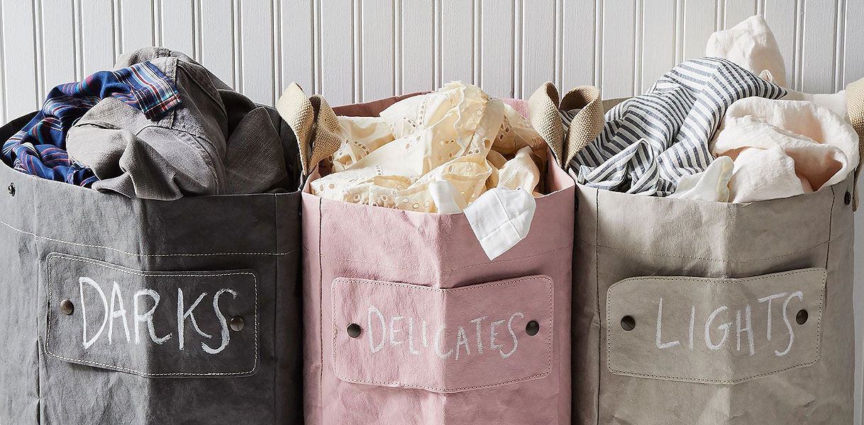 Drie stoffen waszakken in grijs, roze en beige, gelabeld met DARKS, DELICATES en LIGHTS, gevuld met gesorteerde was