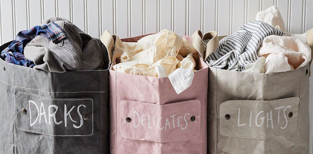 Drie stoffen waszakken in grijs, roze en beige, gelabeld met DARKS, DELICATES en LIGHTS, gevuld met gesorteerde was