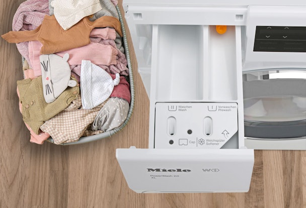 Open wasmiddellade van een Miele wasmachine naast een wasmand met kleding