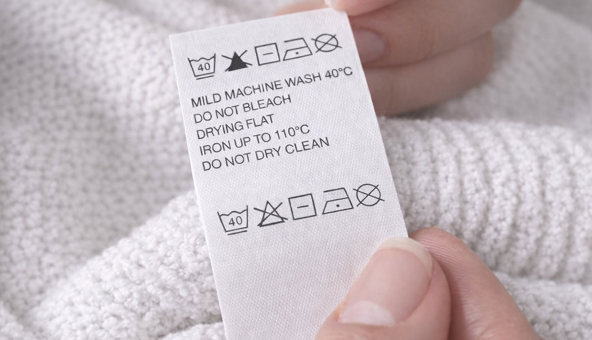 Hand houdt waslabel met wassymbolen vast: machinewas 40 graden, niet bleken, liggend drogen, strijken tot 110 graden