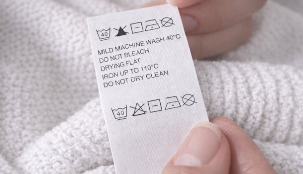 Hand houdt waslabel met wassymbolen vast: machinewas 40 graden, niet bleken, liggend drogen, strijken tot 110 graden