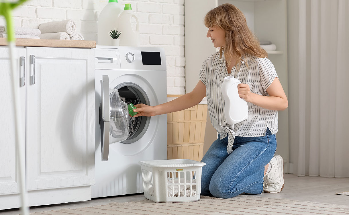 Vrouw knielt voor een wasmachine en stopt wasgoed in de trommel terwijl ze een fles wasmiddel vasthoudt